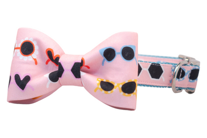 A pink dog collar with an attached bow tie, both printed with colorful novelty sunglasses including daisy-shaped, heart-shaped, hexagonal, and cat-eye designs. The matching dog collar features a silver D-ring, and silver side-release buckle