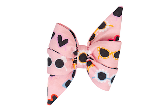 Oversized dog belle bow made from light pink fabric printed with various colorful sunglasses styles, such as black hearts, floral rims, and retro cat-eyes in orange and yellow. The bow has dramatic, sharply angled tails and gathered center folds for a voluminous look.
