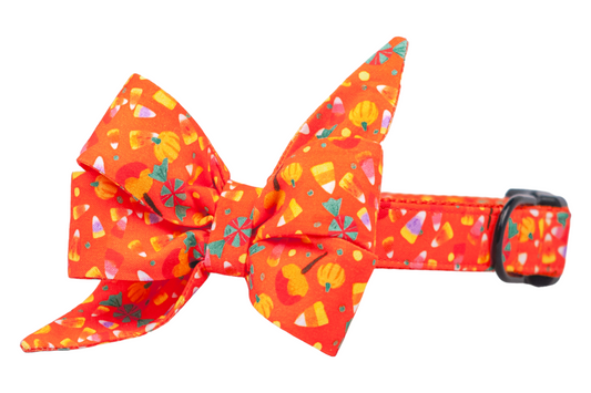Bright orange dog collar with an oversized double-layered belle bow in a fun Halloween print. The design features candy corn, orange pumpkins, peppermint candies, and caramel apples on a vibrant background. The dramatic belle bow with pointed ribbon tails gives this accessory a charming, seasonal look, perfect for festive pet portraits or trick-or-treat outings.