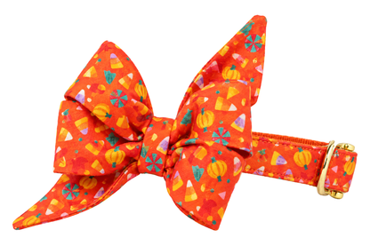Dog collar featuring a large belle bow in Cute and Corny fabric, a bright orange-red print with candy corn, small pumpkins and colorful Halloween candies, attached to a matching Cute and Corny collar with a nylon inner lining and gleaming gold hardware, including a D-ring and buckle.