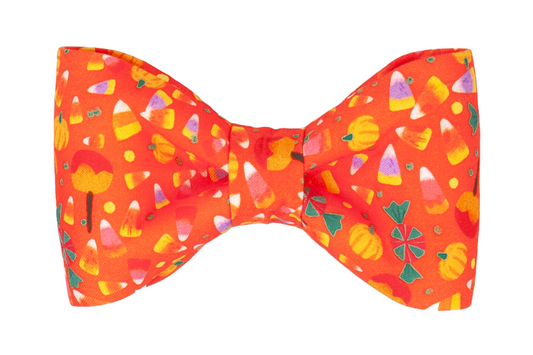A bright orange fabric bow tie featuring a festive Halloween print with scattered illustrations of candy corn, caramel apples, peppermint swirls, and small pumpkins. The bow tie has a structured shape with a gathered cinched center, designed to be worn by dogs as the perfect spooky accessory. 