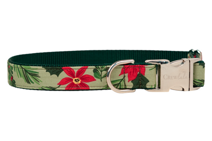 Christmas-themed dog collar with a soft green nylon base and a decorative outer layer with poinsettias, dark green holly leaves, and pine needles. The collar is fitted with polished hardware, including a polished buckle engraved with “CREWlala”.