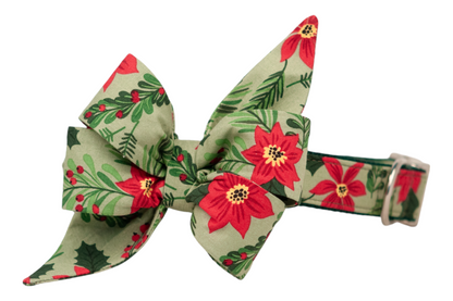 A festive dog collar with an attached oversized belle bow featuring long, pointed tails. Both the collar and bow are made from matching light green fabric decorated with poinsettias, holly leaves, pine sprigs, and berries. The structured belle bow adds a dramatic, holiday-ready flair, while the collar includes hardware for adjustability.