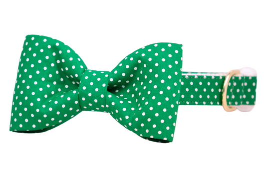 A cheerful green collar for dogs, decorated with small white polka dots and a large matching bow tie. The bow tie is securely attached to the collar, which features durable stitching, a gold D-ring, and a white buckle for easy fastening.