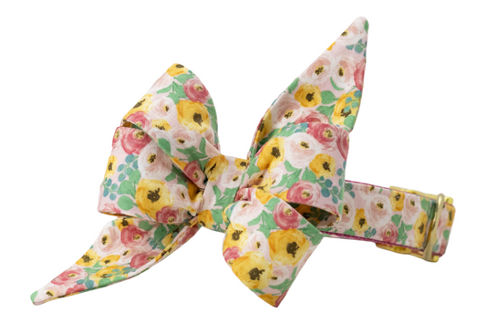 Dog collar with an attached oversized belle bow, both made from cotton fabric featuring a vibrant floral print. The belle bow has an exaggerated structure with two tall, pointed loops and broad lower folds, giving it a sculpted, feminine silhouette. The collar is covered in matching floral fabric and secured with metal hardware, including an adjustment slider and D-ring.