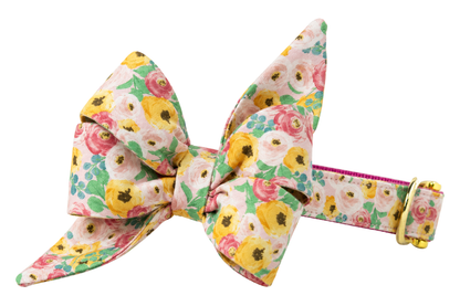 Dog collar featuring a large belle bow in Fairytale Floral fabric, a cream print with painterly yellow, blush pink and white roses and green leaves, attached to a matching Fairytale Floral collar with a nylon inner lining and gleaming gold hardware, including a D-ring and buckle.