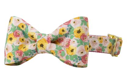 Dog collar with an attached, matching classic bow tie, both made from a soft cotton fabric printed with a vibrant floral pattern. The bow tie has a symmetrical, structured shape with gently curved wings and a neatly cinched center. The collar includes hardware, including a side-release buckle and D-ring.