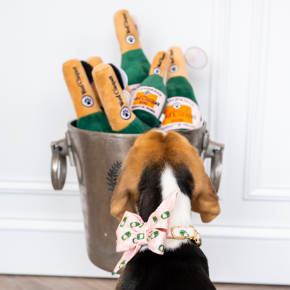 A beagle puppy with a tricolor coat stands on a light wood floor, facing away from the camera and looking into a silver ice bucket filled with plush toy champagne bottles. The dog wears a pink belle bow collar with a champagne bottle and flute pattern, matching the playful theme of the toys.