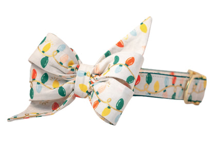 A festive dog collar featuring an oversized, pointed belle bow made from cream-colored fabric printed with colorful string lights. The design includes red, green, yellow, blush, and blue bulbs connected by metallic wires. The collar has an adjustable strap and a polished buckle.