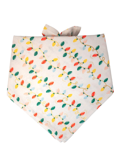 A holiday-themed dog bandana with a cream-colored background and a repeating print of colorful Christmas string lights. The bulbs are red, green, yellow, blue, and peach, connected by looping cords arranged diagonally across the fabric. The bandana ties at the top and has a classic look