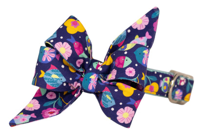 A navy blue dog collar with a large decorative belle bow, featuring a vibrant print of colorful fish, flowers, and polka dots. The matching collar is secured with a metallic buckle.