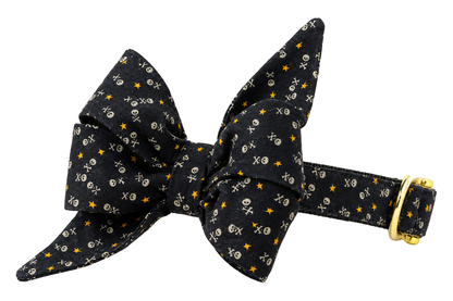 Dog collar featuring a large belle bow in Fright Night fabric, a black fabric with tiny white skulls and crossbones and small orange and yellow dots, attached to a matching Fright Night collar with a nylon inner lining and gleaming gold hardware, including a D-ring and buckle.