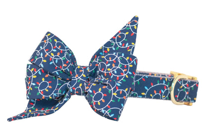 Festive navy blue dog collar featuring an oversized belle bow attachment decorated with strings of multicolored holiday lights. Finished with shiny hardware.