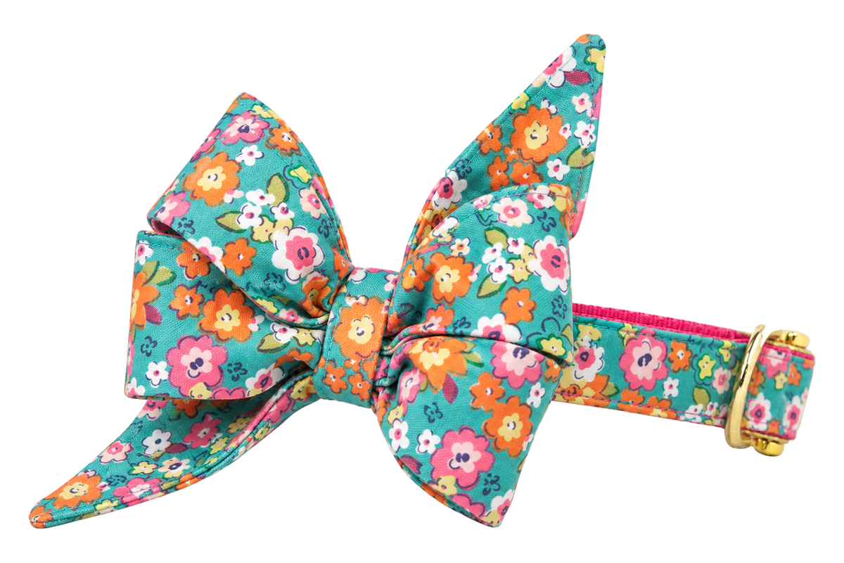 Dog collar featuring a large belle bow in Gigi's Garden fabric, a teal green print with pink, orange, yellow and white flowers, attached to a matching Gigi's Garden collar with a nylon inner lining and gleaming gold hardware, including a D-ring and buckle.