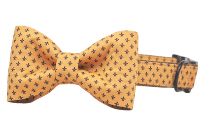 Golden yellow dog collar with a matching oversized bow tie, both adorned with a repeating pattern of small fleur-de-lis motifs. The collar has detailed stitching along the edges and sturdy metal hardware.