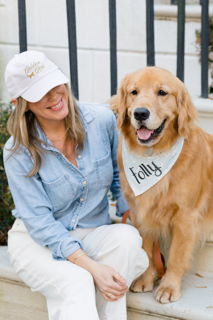 A woman wearing a white “Golden Girl” baseball cap and chambray shirt kneels next to a golden retriever on a stone step. The dog, named Folly per its printed bandana, smiles with its mouth open.