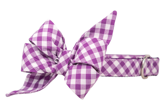 Adjustable dog collar with an attached belle bow in a classic purple plaid pattern. The belle bow has two large loops and two pointed tails, giving it a full, feminine  shape. The collar features visible stitching, a metal D-ring, and is made from sturdy fabric over nylon webbing for durability and comfort.