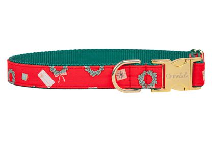 A festive red dog collar decorated with a holiday print of wreaths, wrapped gifts, and tags. The collar is backed with contrasting webbing and features polished hardware, including a D-ring and a side-release buckle engraved with the brand name “Crewlala.”