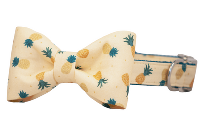 Tropical-themed dog collar with a matching bow tie, decorated with a cheerful pineapple pattern on soft yellow fabric. The pineapples vary in color and orientation, and the silver hardware adds a clean, polished finish to this summery accessory.