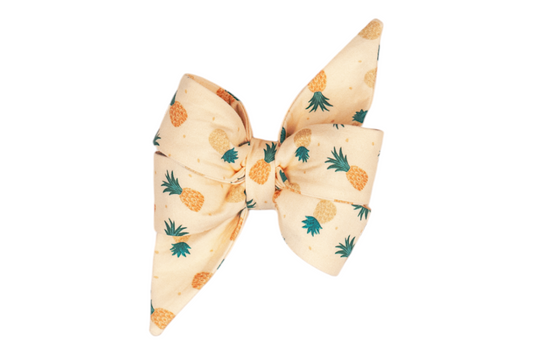 Oversized feminine dog belle bow in a soft yellow color, printed with cheerful pineapples in golden yellow and muted orange, topped with teal-green crowns. The bow is structured with wide folds and dramatic pointed ends, ideal for festive or summer-themed styling.