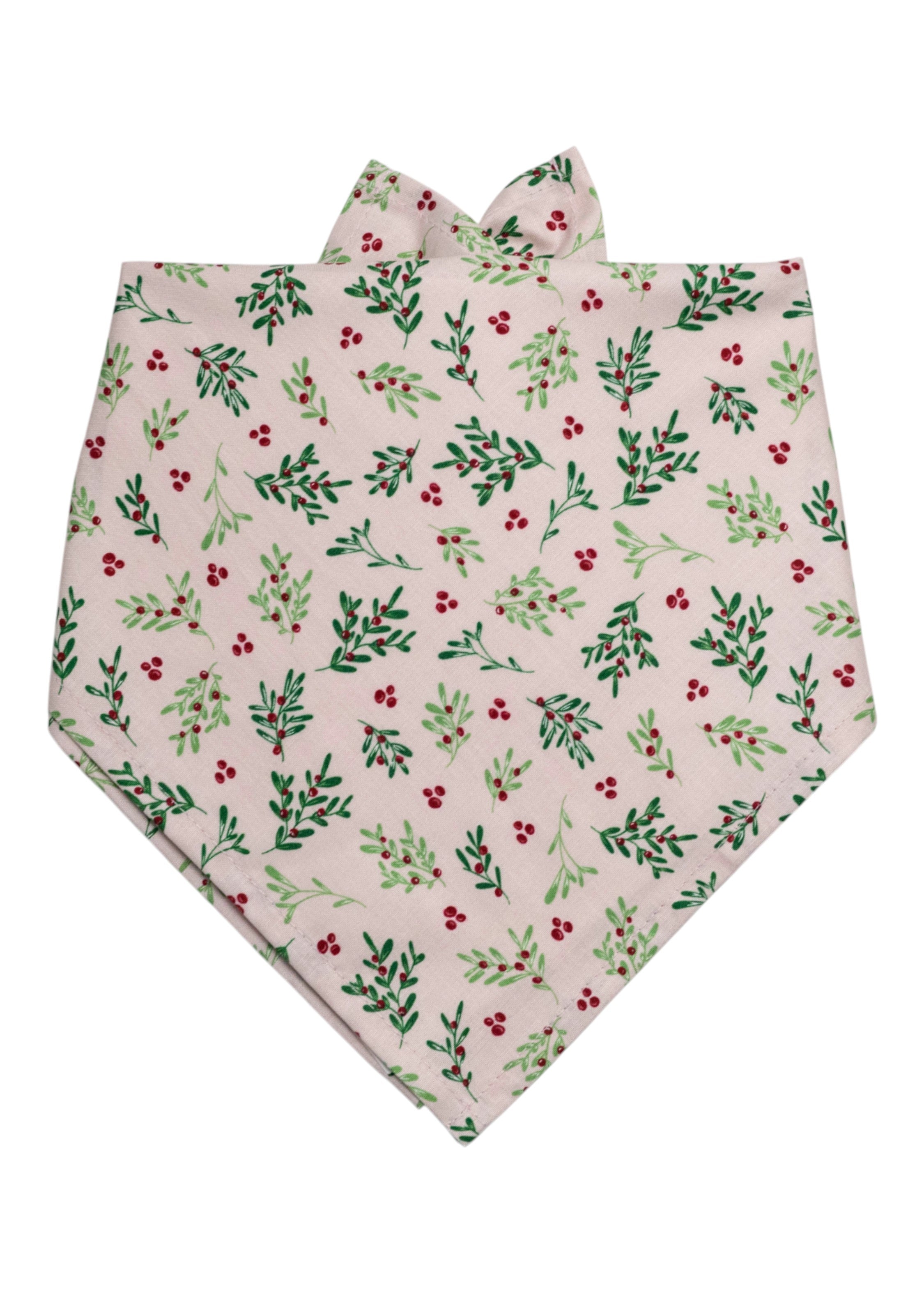 A festive dog bandana in a soft blush pink color, covered in a repeating botanical print of foliage and berries. The triangular fold allows it to be tied comfortably around a dog's neck, with visible folds at the top and a pointed bottom edge that adds a stylish finishing touch.
