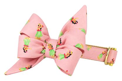 Dog collar featuring a large belle bow in Hula Girls fabric, a soft pink fabric with small hula girls in green grass skirts, attached to a matching Hula Girls collar with a nylon inner lining and gleaming gold hardware, including a D-ring and buckle.