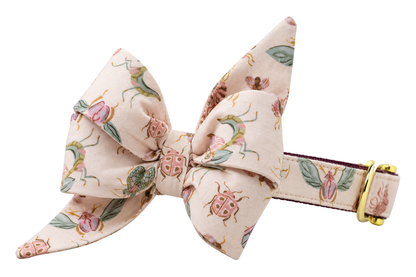 Dog collar featuring a large belle bow in Love Bugs fabric, a soft blush cream fabric with vintage-style beetles, moths and green leaves, attached to a matching Love Bugs collar with a nylon inner lining and gleaming gold hardware, including a D-ring and buckle.