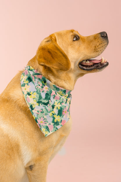 A happy golden Labrador sits upright, facing to the right with ears perked and teeth showing. Around its neck is a soft bandana printed with green, pink, and yellow watercolor-style flowers. The background is a warm, even-toned pink, highlighting the dog’s shiny coat and colorful accessory.
