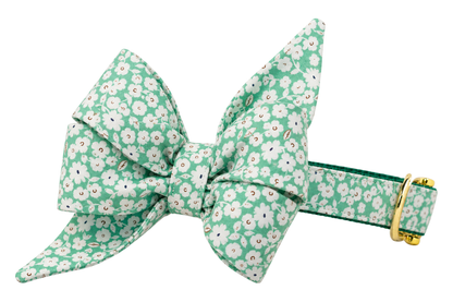 Dog collar featuring a large belle bow in Lucky Meadow fabric, a green fabric with a dense pattern of small white clover flowers, attached to a matching Lucky Meadow collar with a nylon inner lining and gleaming gold hardware, including a D-ring and buckle.