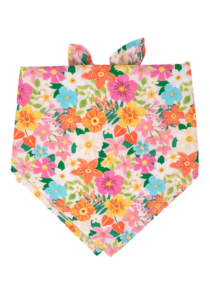 Vivid floral dog bandana folded into a symmetrical triangle shape, covered in a dense pattern of tropical-style flowers in sunny shades of orange, magenta, turquoise, lemon yellow, and white. Green foliage and delicate pink buds fill the background, creating a lively, summer-inspired look.