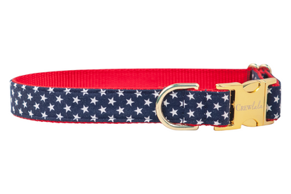 Close-up of a patriotic dog collar made from star-patterned navy fabric with a red woven interior. The collar is finished with polished gold-toned metal, including a branded clasp and sturdy D-ring for leash attachment.
