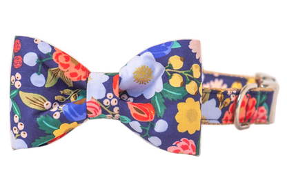 Dog collar with a matching attached bow tie, featuring a navy blue background with a lively floral pattern and berries. The bow tie is centered on the collar, which has nylon webbing and a metal D-ring.
