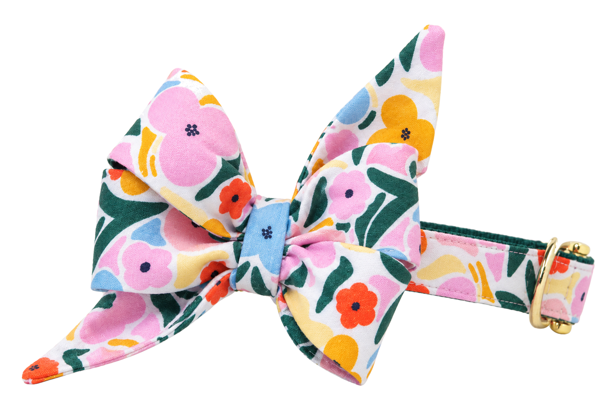 Dog collar featuring a large belle bow in Mosaic Blooms fabric, a white print with bold pink, sky blue, yellow and red-orange flowers and dark green leaves, attached to a matching Mosaic Blooms collar with a nylon inner lining and gleaming gold hardware, including a D-ring and buckle.