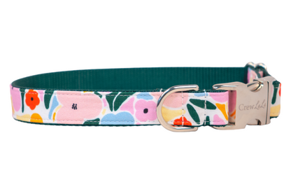 A stylish dog collar featuring a colorful floral print with pink, orange, yellow, and blue flowers and green leaves on a white background. The collar has a dark green inner lining and is finished with a shiny silver metal buckle and D-ring. The brand name “Crewlala” is engraved on the buckle.