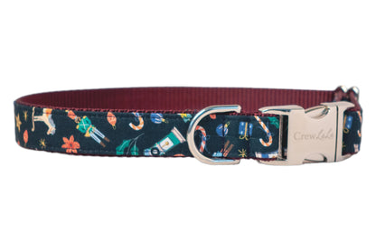 A festive dog collar with a navy blue outer layer patterned with colorful holiday illustrations—nutcrackers, rocking horses, candy canes, holly, and presents—set against a sturdy nylon webbing lining. It includes a metal D-ring for leash attachment and a polished buckle clasp engraved with “Crew Lala.”