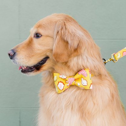 Side view of a golden retriever wearing a matching yellow leash and bow tie set, printed with fun oyster, pearl, lemon, and oyster knives illustration, in front of a green brick wall.