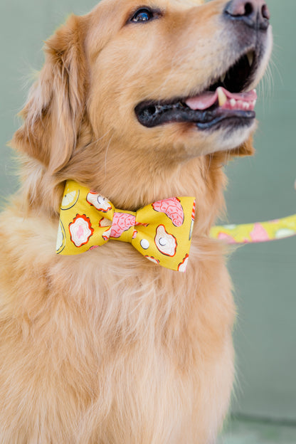 Close-up of a golden retriever’s face and chest, showing a cheerful yellow bow tie with a oyster print that includes oysters, pearls, lemons, and knives in pink, white, and orange. The matching leash is visible behind the dog’s neck.