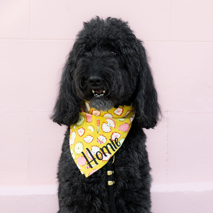 A large, black, curly-coated dog with an open mouth sits calmly against a pink wall, wearing a vivid yellow bandana with oysters, pearls, lemons, and oyster knives print. Embroidered in the corner is “Homie” in script.