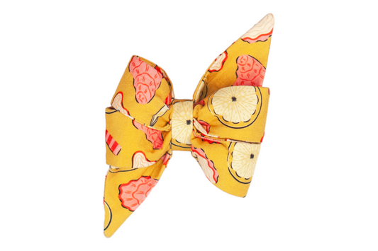 Fabric feminine dog belle bow with a summery yellow background, printed with whimsical illustrations of oysters, lemon slices, pearls, and oyster knives in coral, cream, and black accents. The bow is large and structured, with exaggerated pointed tails.