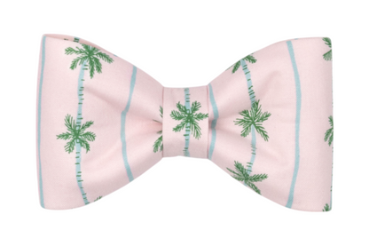 A standalone dog bow tie in pastel pink, printed with green palm trees and light blue vertical stripes. The tropical bow tie is symmetrical and pre-tied.