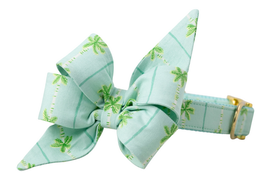 Tropical-themed dog collar in an aqua blue with a matching oversized bow, both printed with green palm trees and aqua vertical lines. Features shiny gold hardware and teal inner webbing.