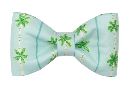 Tropical-themed dog bow tie in aqua blue, decorated with a repeating green palm tree and blue stripe pattern. Designed for collar attachment.