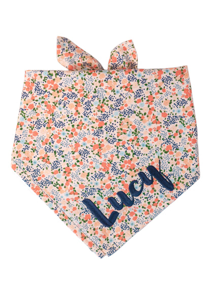 Dog bandana in pale peachy pink fabric, covered in a dense floral print with green foliage, designed to tie around the neck. The name "Lucy" is embroidered on the right side.