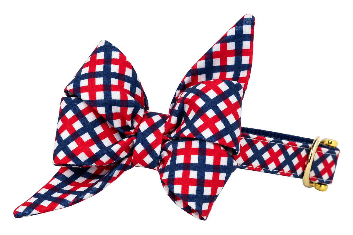 Dog collar featuring a large belle bow in Patriotic Plaid fabric, a bold red, navy and white lattice plaid, attached to a matching Patriotic Plaid collar with a nylon inner lining and gleaming gold hardware, including a D-ring and buckle.