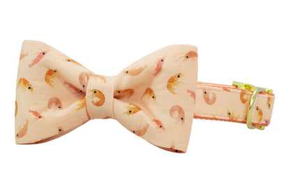 Peach-colored dog bow tie collar with a repeating shrimp pattern in soft pink and orange tones, attached to a matching adjustable collar with gold hardware.