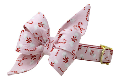A holiday dog collar with an oversized belle bow attachment, both made from soft pink fabric printed with candy canes and peppermint swirls. The bow features large, structured loops and long pointed tails, while the collar is backed in nylon and finished with polished hardware for festive flair.