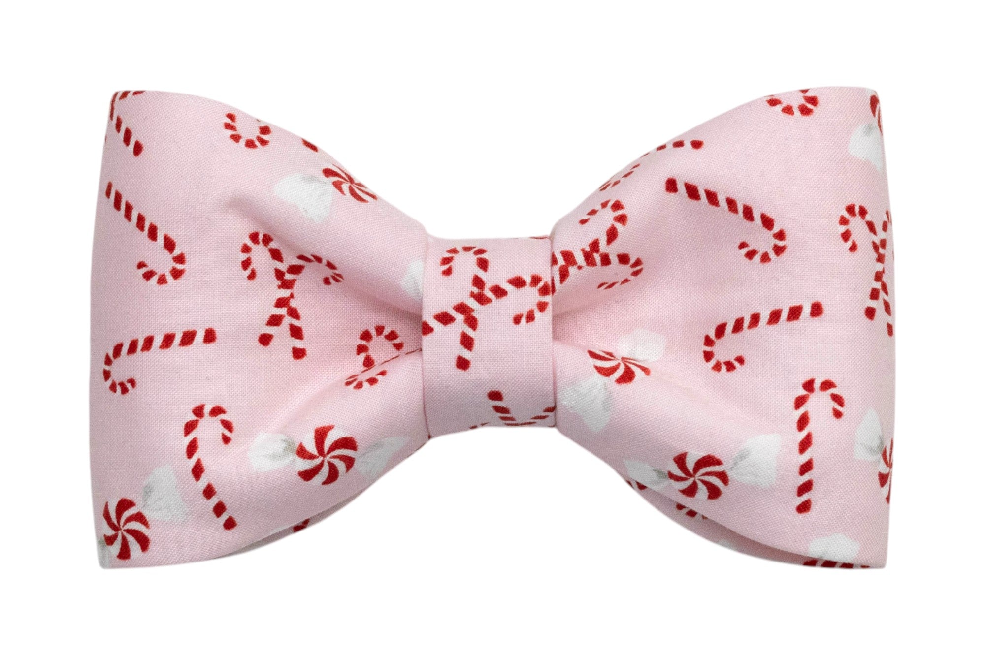 A festive dog bow tie made from light pink fabric printed with peppermint swirls and candy canes. The bow has a structured, symmetrical shape with a cinched center and is designed to attach to a collar for a playful holiday look.