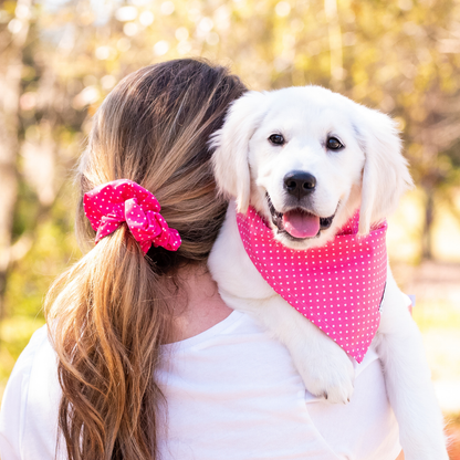 A woman with long blonde hair tied back with a pink polka dot scrunchie holds a smiling white puppy over her shoulder. The puppy wears a matching pink polka dot bandana, and both are outdoors in a sunlit, tree-filled setting.