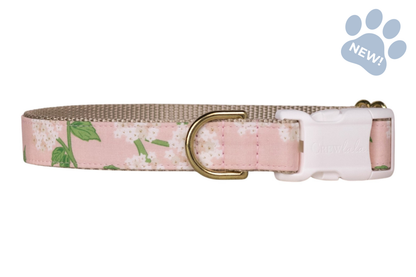 Adjustable dog collar featuring a blush pink hydrangea print with white flower clusters and green leaves, accented by beige webbing and gold-tone hardware and a sturdy white plastic buckle.