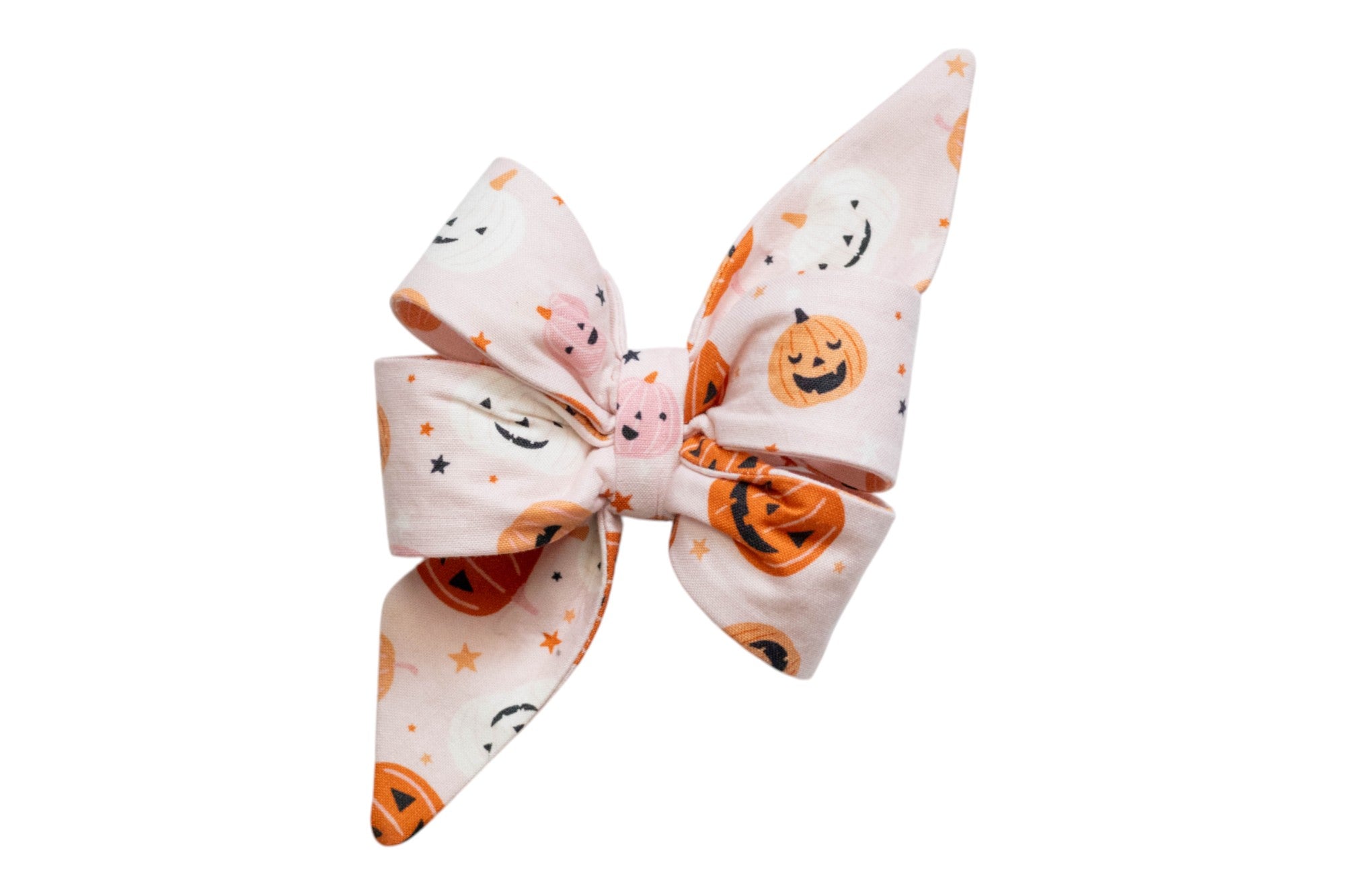 Halloween-themed dog bow with a soft pink base and oversized, layered shape including two long tails. The fabric has smiling pumpkins in a mix of tones. Each pumpkin has a carved face, ranging from cute to spooky, surrounded by scattered five-pointed stars in coordinating Halloween colors.